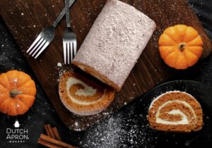 Pumpkin Spice Season Specialty - Pumpkin cake roll with a slice taken out and another slice next to it. 2 forks and 2 pumpkins are next to it on a cutting board with cinnamon sticks and powdered sugar for garnish.