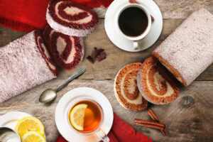 Drink Pairings - Partially cut red velvet and pumpkin cake rolls with 2 white cups and saucers filled with coffee and tea. Spoons, lemons, and cinnamon sticks for garnish on wooden tabletop.
