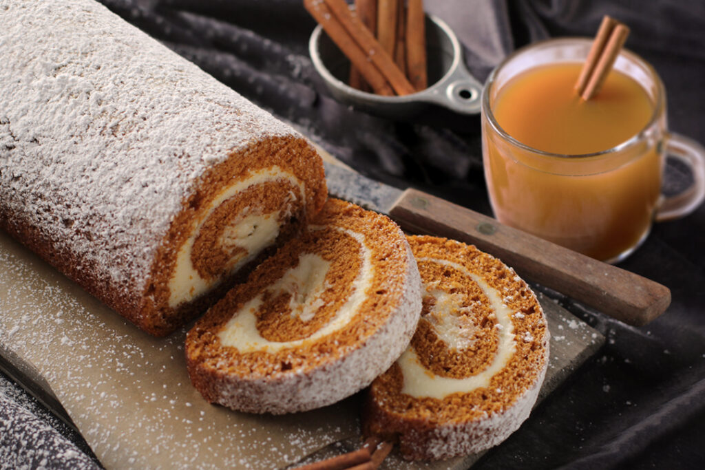 Drink pairings - Partially cut pumpkin cake roll on cutting board with knife and a cup of cinnamon sticks next to it along with a clear mug of cider with cinnamon sticks inside.