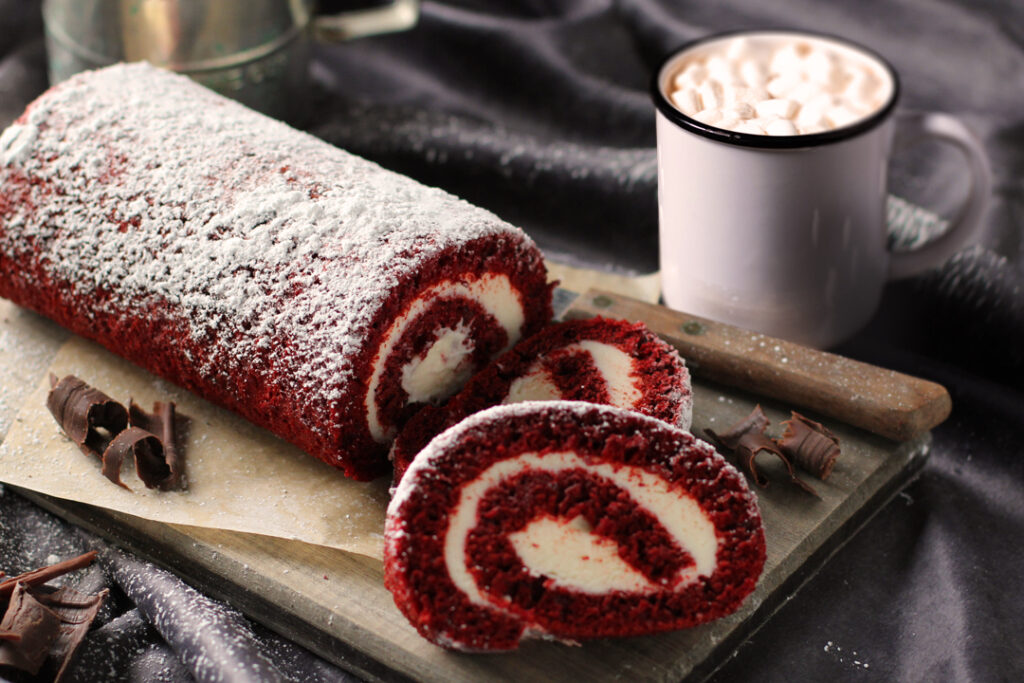 Partially cut red velvet cake roll with chocolate shavings as garnish on cutitng board with knife. Mug of hot chocolate with marshmallows next to it.