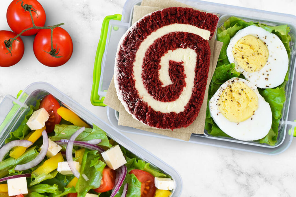 Slice of Red Velvet Cake Roll in school lunch box next to hard-boiled egg, salad, tomatoes, and more.