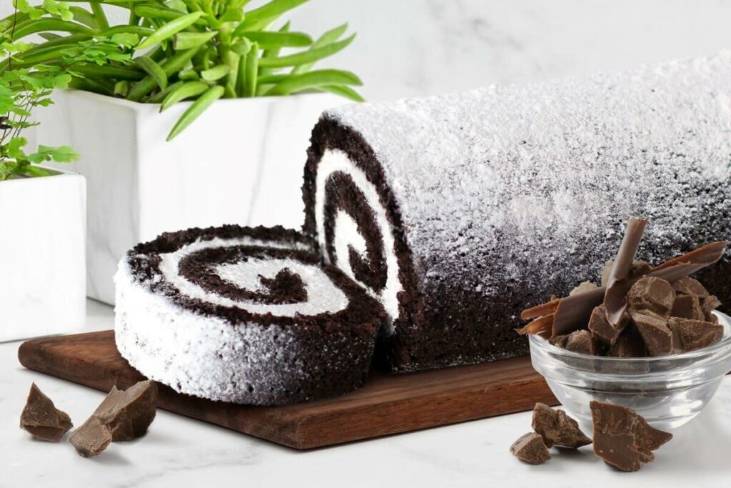 Chocolate Crème Cake Roll on serving platter next to cup of chocolate pieces with a plant in the background.