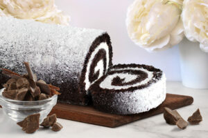 Spring Cake Flavors: Chocolate Crème Cake Roll with slice on wooden platter with white flowers in background and chocolate pieces in the front.