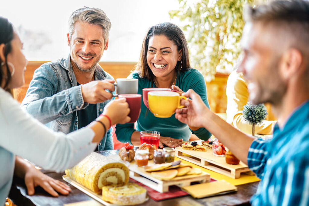 Tips for Hosting Brunch main blog image - people sitting around an outside table, clinking their coffee mugs together, with breakfast foods and treats on the table in front of them.