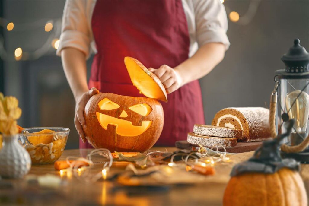 Woman carving a pumpkin on a table filled with decorations, pumpkin carving tools, and a pumpkin cake roll.