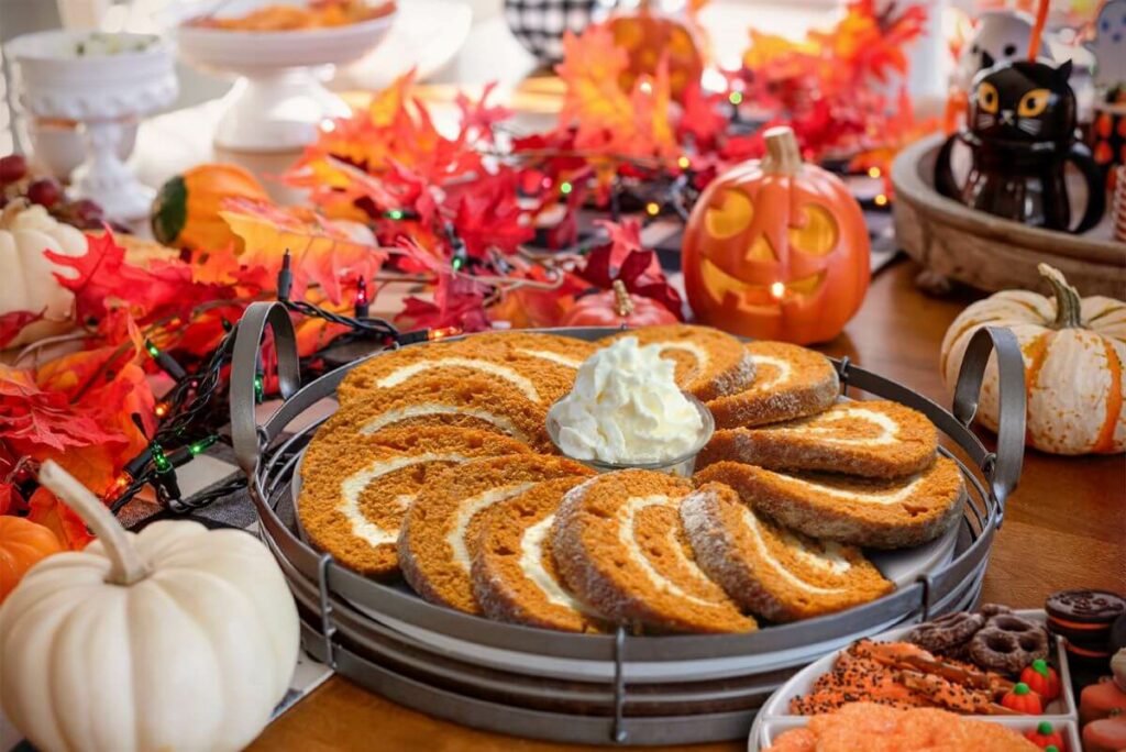 Series of Pumpkin Cake Roll slices on a serving platter. Table is decorated with fall and Halloween decorations for the Pumpkin Carving Party. There are also other treats on the table.
