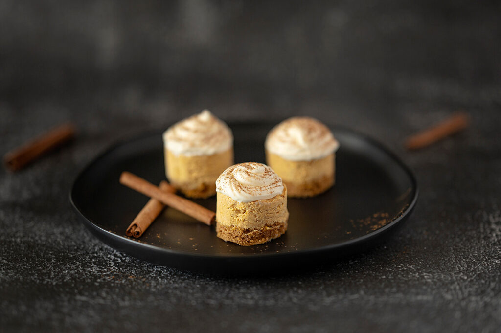 Close up of one Mini Pumpkin Cheesecake on a plate with two others in the back. Cinnamon sticks are also on the plate.