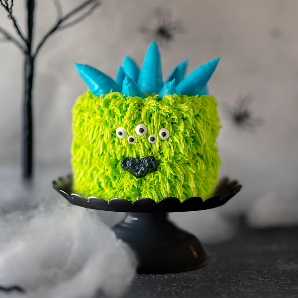 Green and blue Mini Monster Cake Roll on cake pedestals with a background of spider webs, fake spiders, and ghoulish trees.