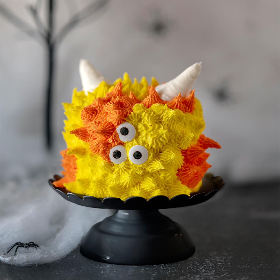 Orange and yellow Mini Monster Cake Roll on cake pedestals with a background of spider webs, fake spiders, and ghoulish trees.