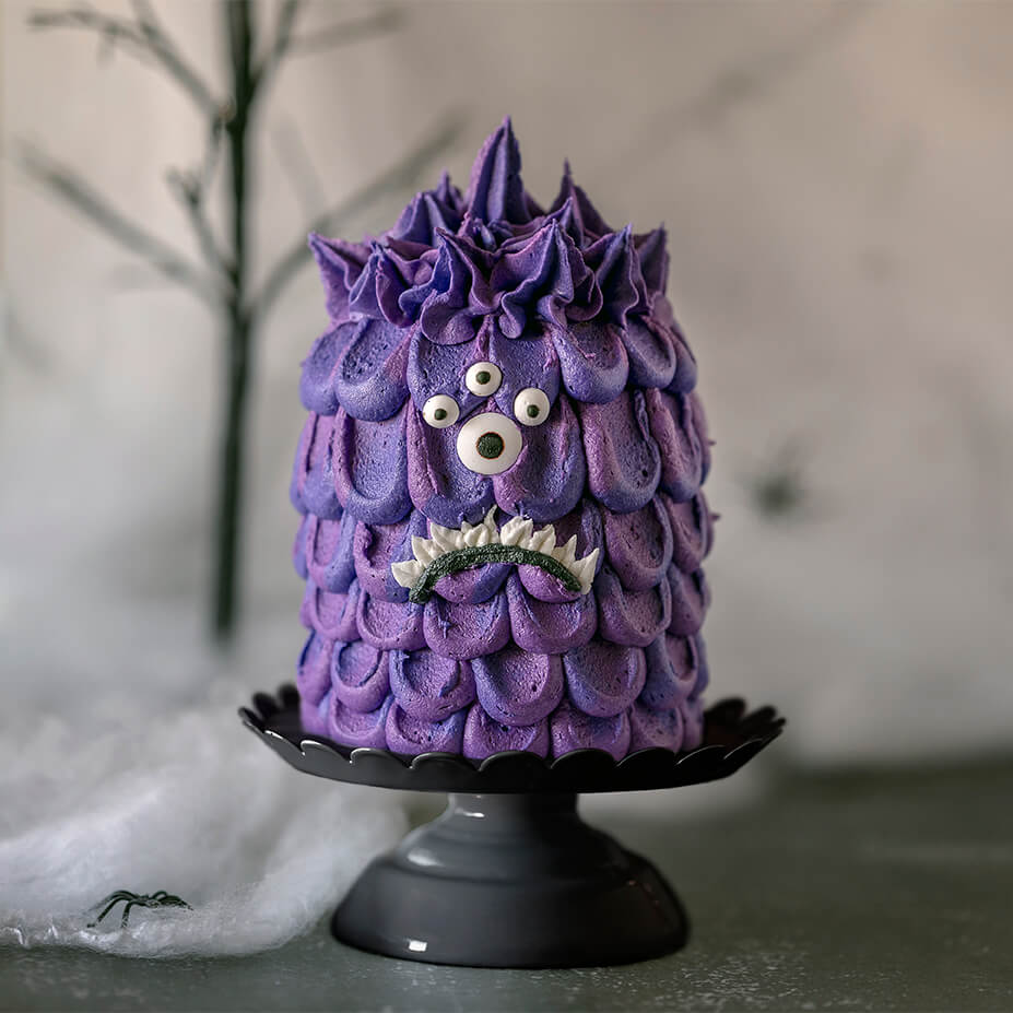 Purple Mini Monster Cake Roll on cake pedestals with a background of spider webs, fake spiders, and ghoulish trees.