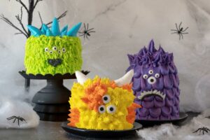 All three Mini Monster Cake Rolls on plates/cake pedestals with a background of spider webs, fake spiders, and ghoulish trees.