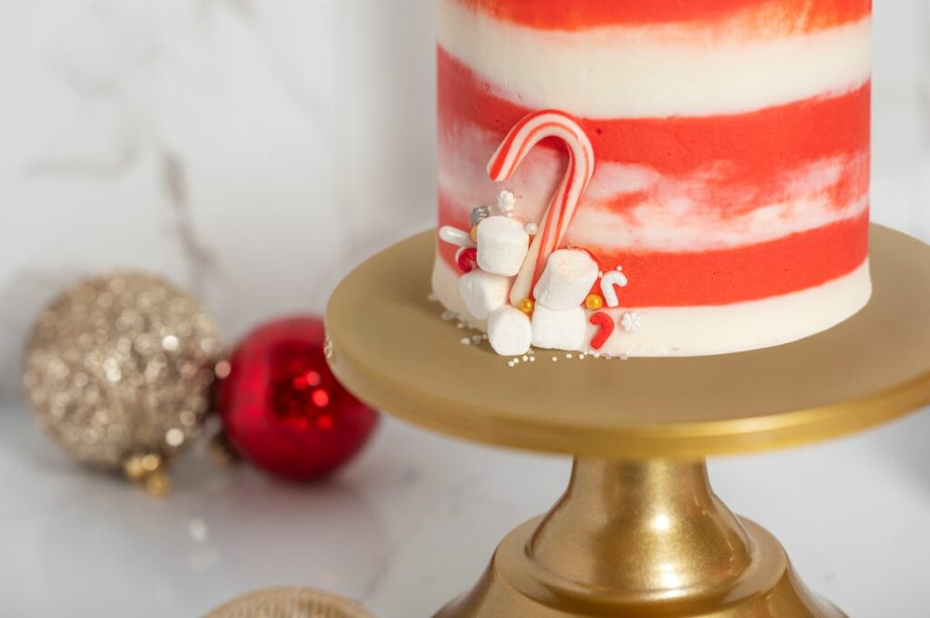 Close up of Winter Wonderland Cake Roll decorations - mini candy cane, sprinkles, and marshmallows. The cake roll is on a gold cake stand and surrounded by red and gold Christmas bulbs.