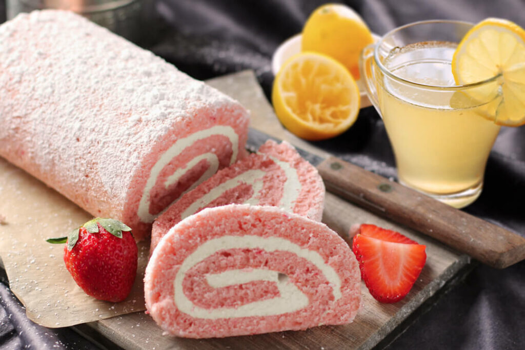 Strawberry Cheesecake Cake Roll on cutting board with real strawberries next to it. It's next to a glass of lemonade.