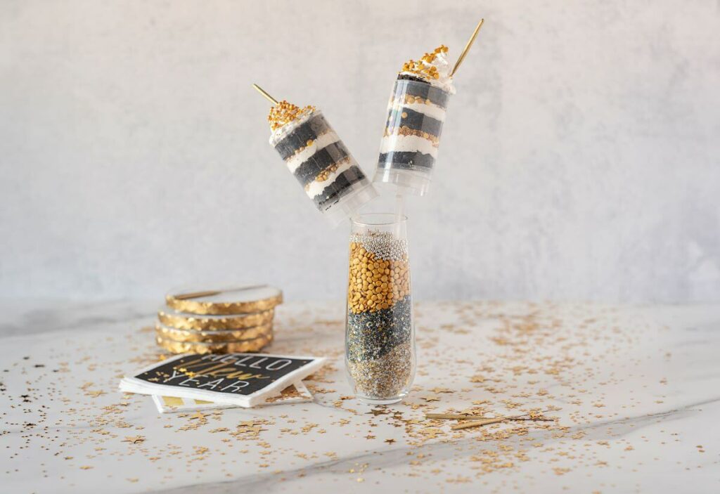 New Year Cake Push Pops with sparklers unlit in a vase. Sprinkles are scattered on the table. The dessert is next to themed napkins and coasters.