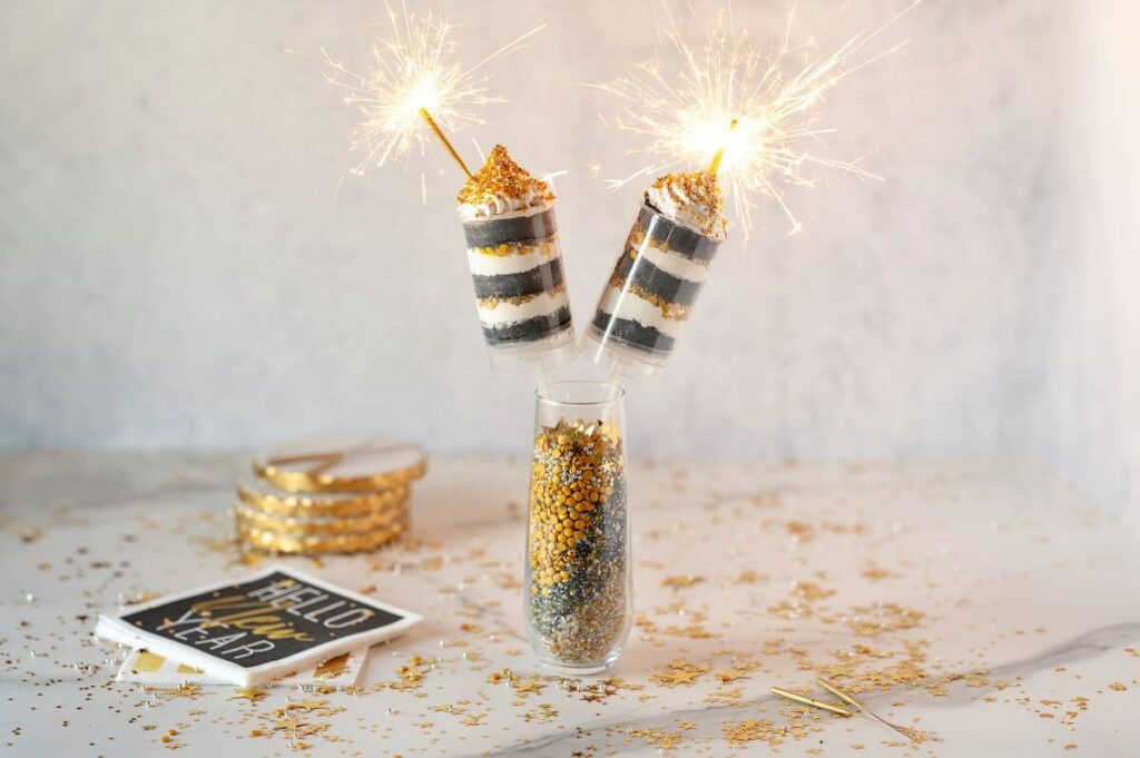 New Year Cake Push Pops with sparklers lit in a vase. Sprinkles are scattered on the table. The dessert is next to themed napkins and coasters.