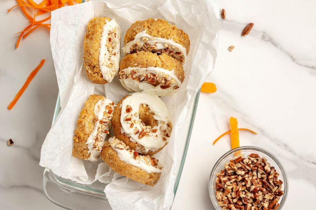 Six Mini Carrot Cake Donuts in a container, ready to serve or take to a gathering. There is a bowl of nuts and carrot shavings around the container.