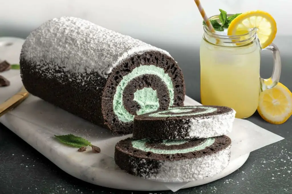 Mint Chocolate Cake Roll on marble cutting board. There are two slices in front of the full cake roll. There's mint leaves and chocolate pieces around the roll. Next to the roll is a mason jar filled with mint lemonade.
