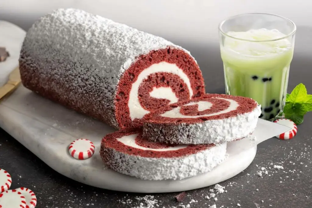 Peppermint Cheesecake Cake Roll on marble cutting board. There are two slices in front of the full cake roll. There's peppermint candies scattered around the roll. Next to the roll is a glass of Matcha Bubble Tea.