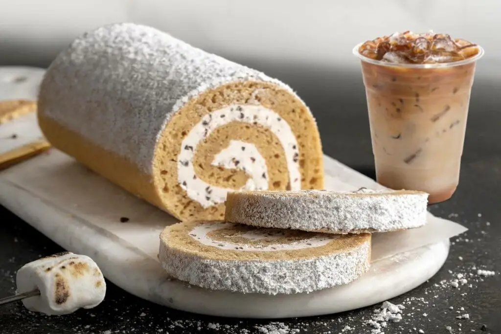 S'mores Cake Roll on marble cutting board. There are two slices in front of the full cake roll. There's a toasted marshmallow next to the cake roll. On the other side of the roll is a glass of Caramel Iced Coffee.