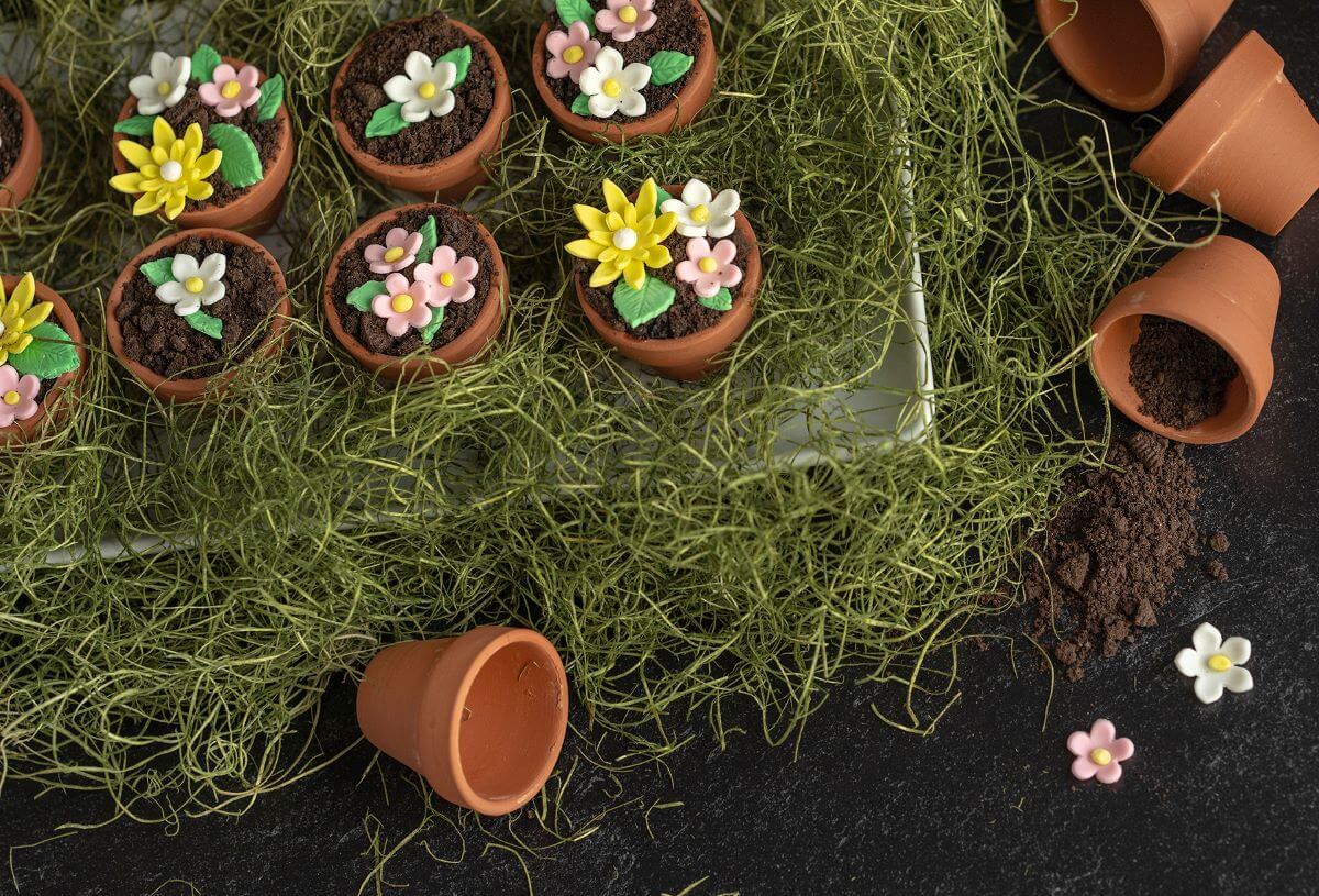 Flowerpot Cakes on a bed of fake grass with several empty flowerpots laying around them, spilling cookie crumb "dirt" and gum paste flowers.