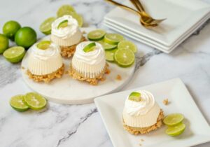 One Mini No-Bake Key Lime Pie on a plate with three more on a serving platter behind it. There's a stack of plates and forks in the background. Key lime slices are used as garnish.