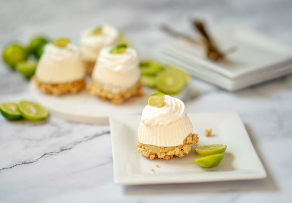 One Mini No-Bake Key Lime Pie on a plate with three more on a serving platter behind it that are out of focus. There's a stack of plates and forks in the background. Key lime slices are used as garnish.