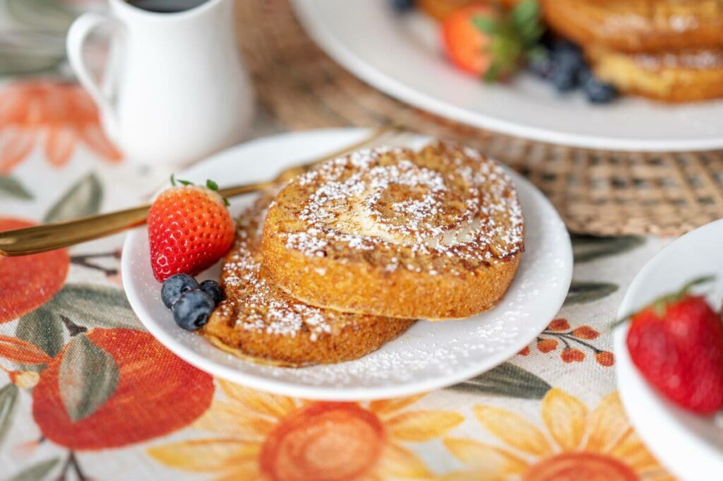 Two slices of Carrot Cake French Toast on a plate. They're covered in powdered sugar and have fresh fruit next to them.