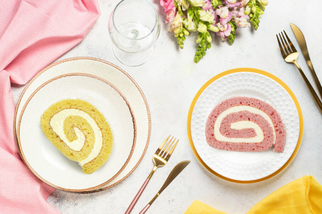 A picture depicting two different place setting options for a Mother's Day Luncheon. One has two stacked plates that are off white with a Lemon Crème Cake Roll in the center of the top plate. The other is a white plate with a gold border and decorative pattern. There's a Strawberry Cheesecake Cake Roll slice on the center. There's also pink and yellow napkins and flower options.