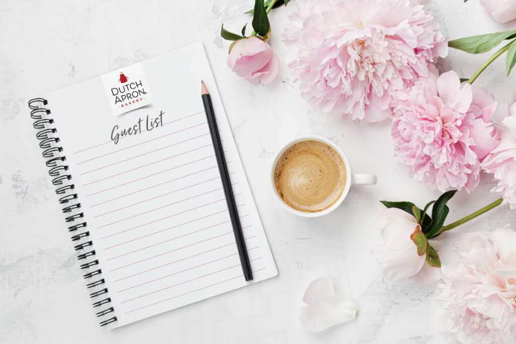 A notepad on a white marble background. It's towards the left of the image. On the right are pink flowers and a cup of coffee. On the notepad, there's the Dutch Apron Bakery logo and "Guest List" written at the top.