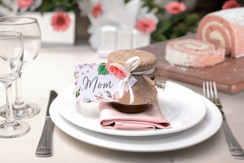 A small jar of jam with a bow wrapped around it and a tag that reads "Mom". It's sitting on a place setting of white plates and a pink napkin. In the background, there are pink flowers and a Strawberry Cheesecake Cake Roll.