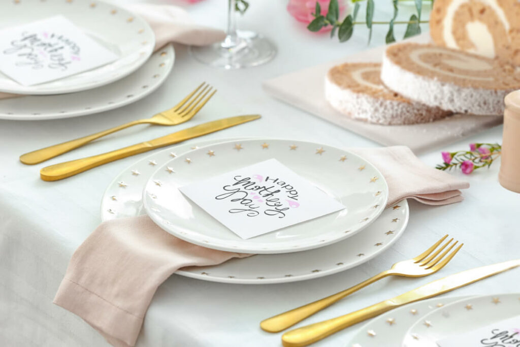 A table set for a Mother's Day Luncheon. There's white plates decorated with gold stars. Each place setting has two of these plates, a pink napkin, and gold silverware. There's a Carrot Cake Cake Roll in the background.