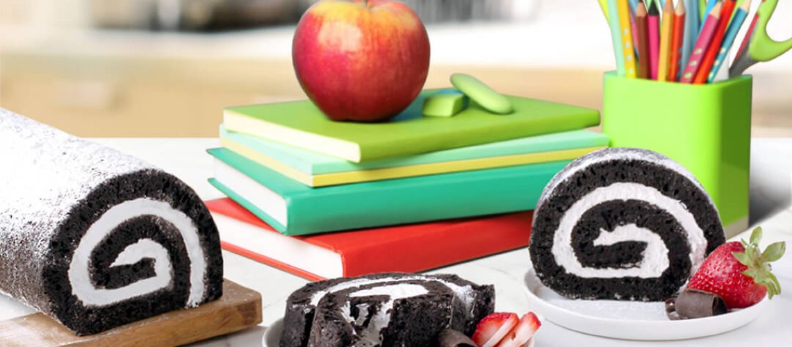 Back to School Treats - Full chocolate cake roll with two chocolate cake roll slices on plates with garnish. On a table next to school books, pencil holder, etc.