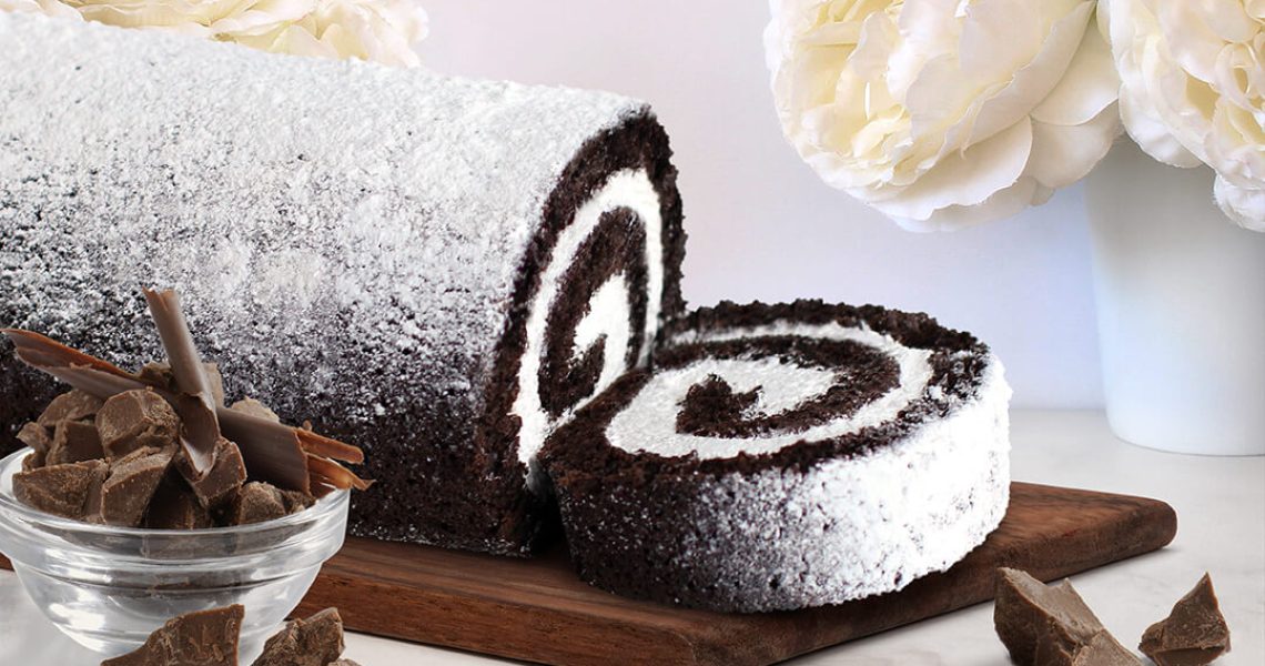 Spring Cake Flavors: Chocolate Crème Cake Roll with slice on wooden platter with white flowers in background and chocolate pieces in the front.