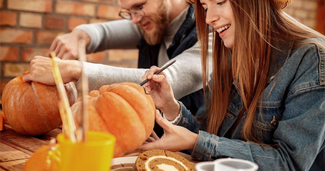 Two people carving pumpkins at at able. On the table are various pumpkin carving tools and slices of a Pumpkin Cake Roll.