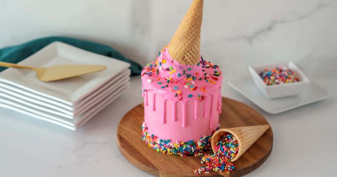 Dripping Ice Cream Cone Cake Roll on serving platter with an ice cream cone full of sprinkles next to it, a bowl of sprinkles in the back, and a pile of plates with a serving spatula to the left.
