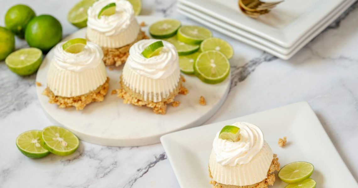 One Mini No-Bake Key Lime Pie on a plate with three more on a serving platter behind it. There's a stack of plates and forks in the background. Key lime slices are used as garnish.