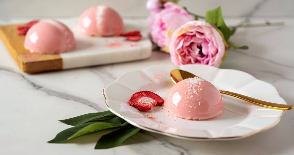 One Mini Strawberry Dome Cake on a plate next to a spoon. There's a strawberries piece and strawberry powder on the plate as well. There are two full dome cakes in the background as well as pink flowers.