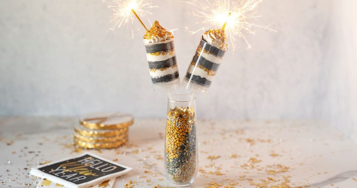 New Year Cake Push Pops with sparklers lit in a vase. Sprinkles are scattered on the table. The dessert is next to themed napkins and coasters.