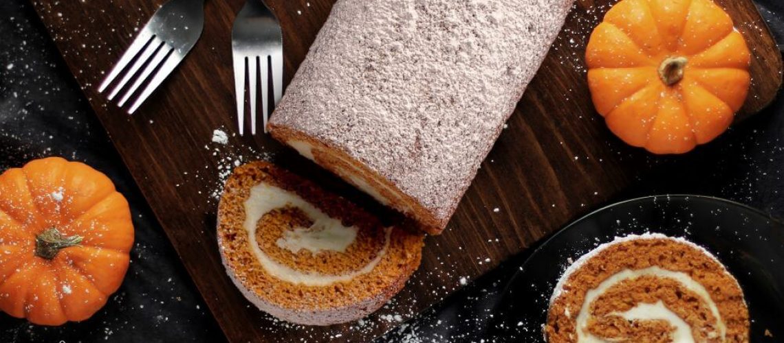 Pumpkin Spice Season Specialty - Pumpkin cake roll with a slice taken out and another slice next to it. 2 forks and 2 pumpkins are next to it on a cutting board with cinnamon sticks and powdered sugar for garnish.